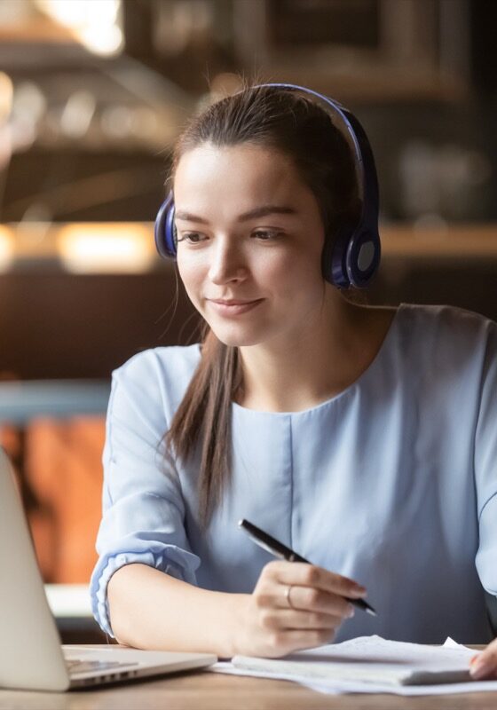 Focused woman wearing headphones using laptop in cafe, writing notes, attractive female student learning language, watching online webinar, listening audio course, e-learning education concept