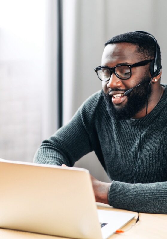 Young African-American guy is call center worker or support. Smart black man in eyeglasses uses a handsfree headset and laptop to talk online at his workplace